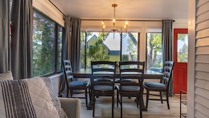A dining area with a table and chairs, large windows with curtains, and a view of a house and trees. - Steps to Restaurants, Shops & Waterfront, Bar Island, Free 2-car Parking,  Wi-fi (Bar Harbor)