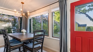 A dining area with a wooden table and chairs, large windows with curtains, and a red door. - Steps to Restaurants, Shops & Waterfront, Bar Island, Free 2-car Parking,  Wi-fi (Bar Harbor)
