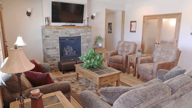 A living room with a stone fireplace, leather sofas, a coffee table, and a television mounted above the fireplace. - Greystone Crossing Suite #8 (Heyburn)