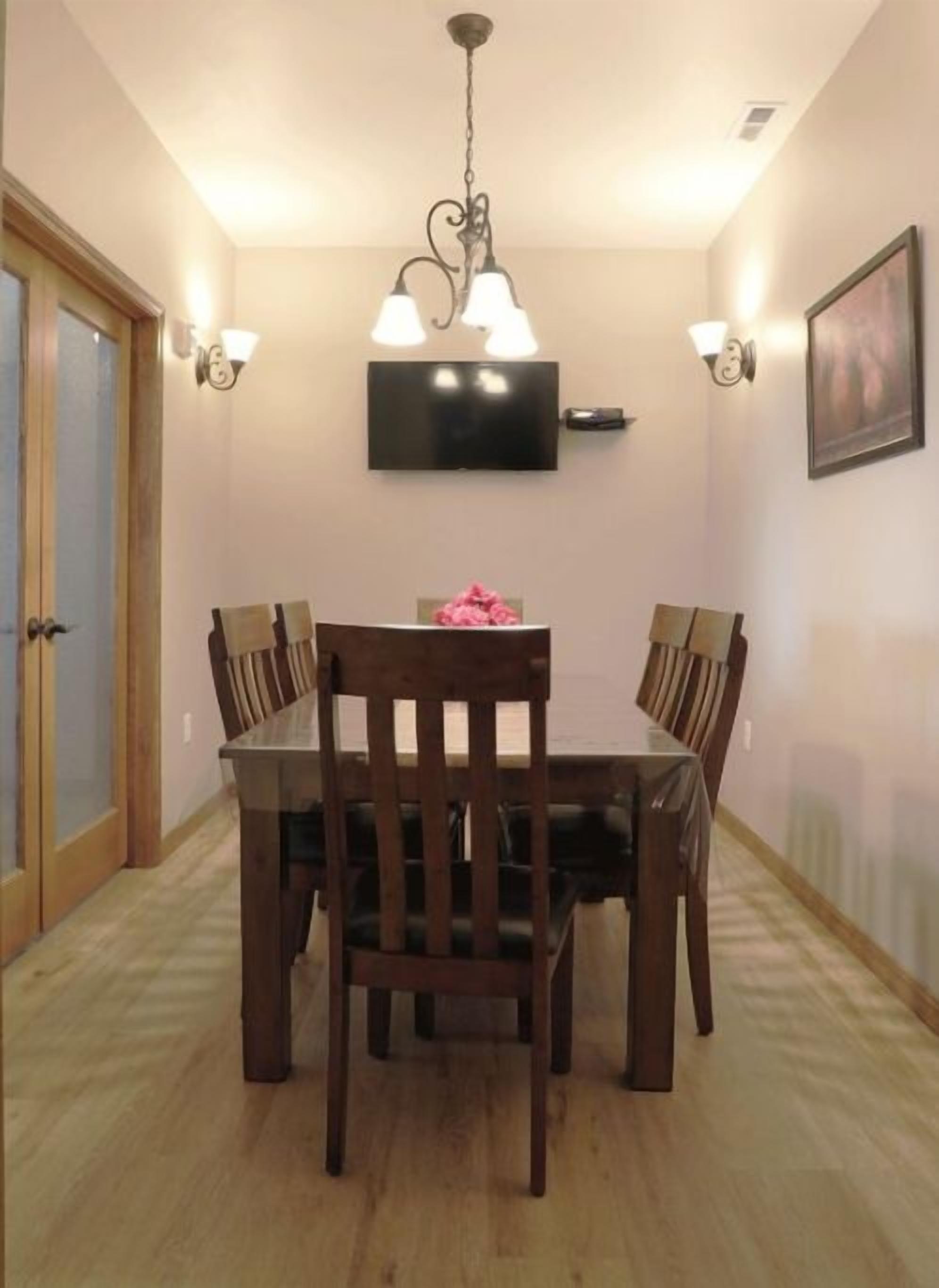 A dining area with a wooden table and chairs, a chandelier, wall-mounted lights, and a television on the wall.