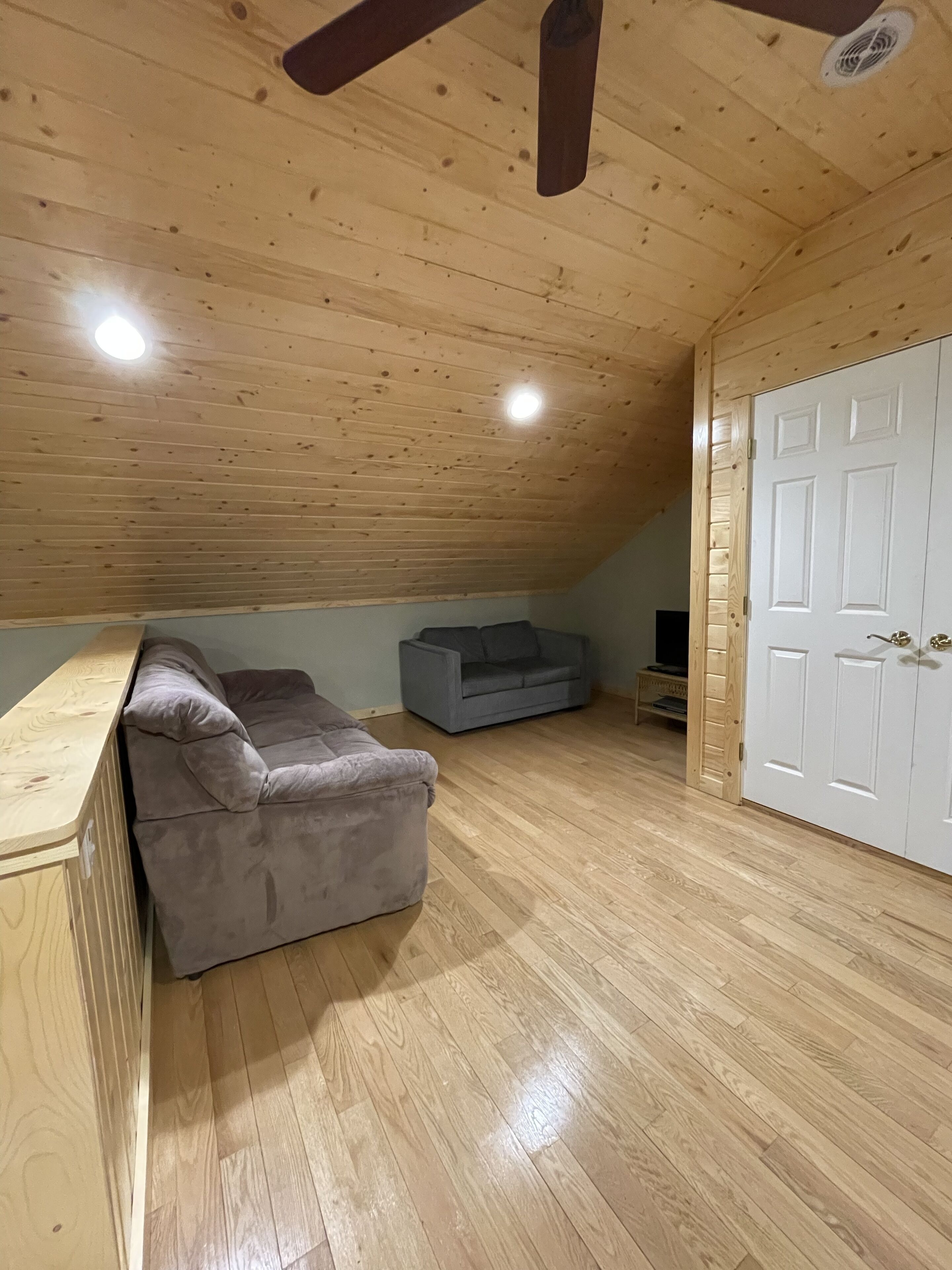 Une pièce de style loft avec un plafond en pente, un plancher en bois, un canapé gris, une petite table et une armoire blanche.