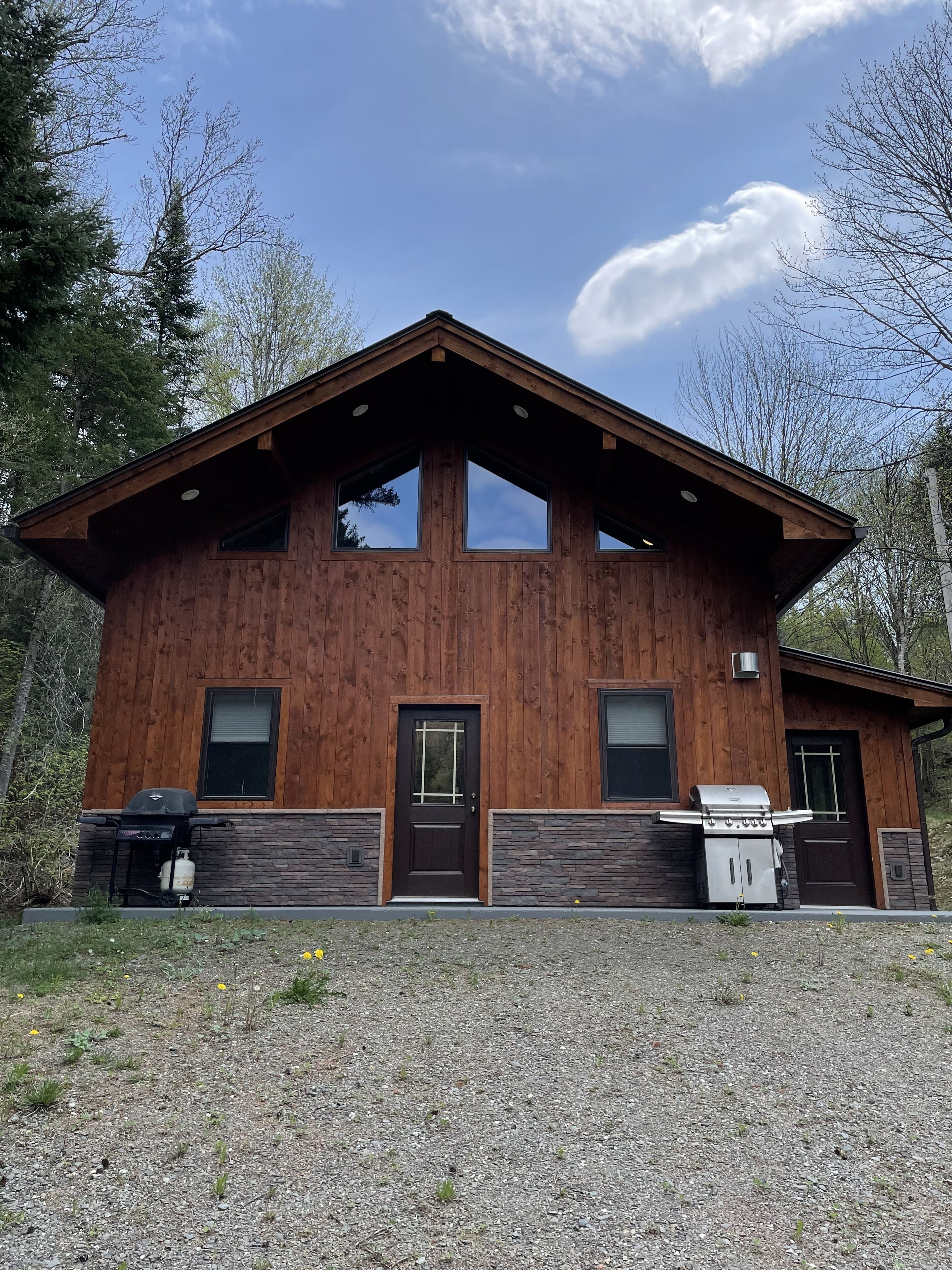 Une cabane en bois avec des fondations en pierre, de grandes fenêtres et un espace barbecue.