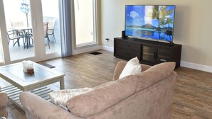 Ein Wohnzimmer mit Fernseher, einer Couch, einem Couchtisch und Blick ins Freie durch eine Glasschiebetür.
