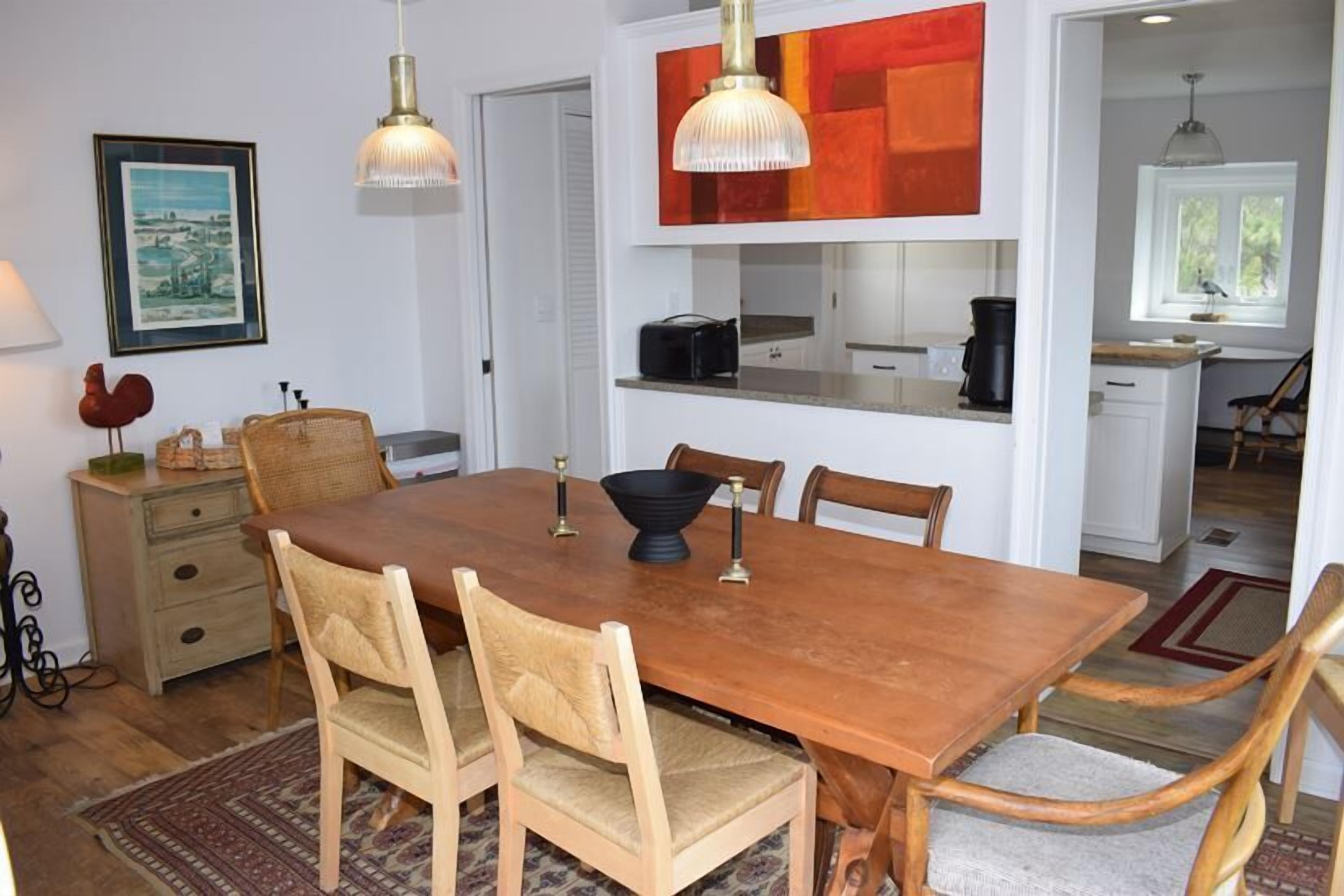 A dining room with a wooden table, six chairs, and a chandelier. There is a kitchen area visible in the background.