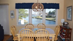 Uma sala de jantar com uma mesa posta para quatro pessoas, cadeiras de madeira, um lustre e vista para a água através de grandes janelas.