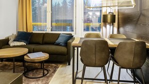 A modern living room with a sofa, coffee table, and chairs. There is a dining table with chairs, a lamp, and a view through large windows.