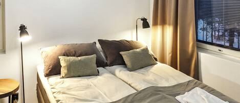 A hotel room with a bed, two bedside lamps, a window with curtains, and a view of trees.