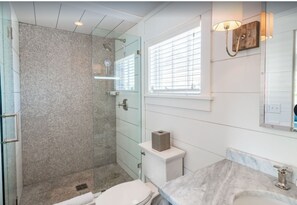 Et badeværelse med glasbrusekabine, toilet, vask med marmorflade og et vindue med persienner.