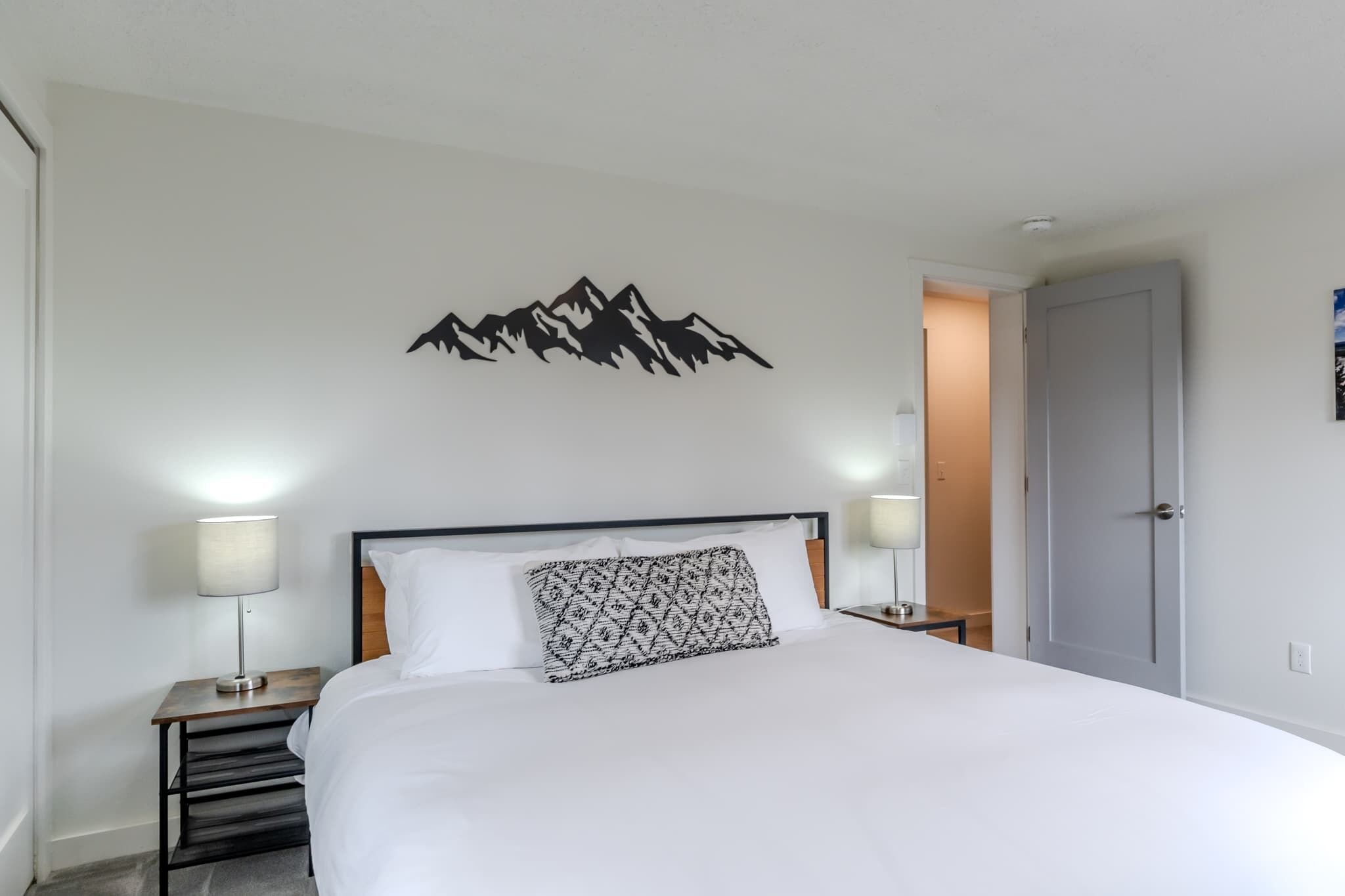Een net opgemaakt bed met een kussen met dessin, twee bedlampen en een wanddecoratie van een berg.