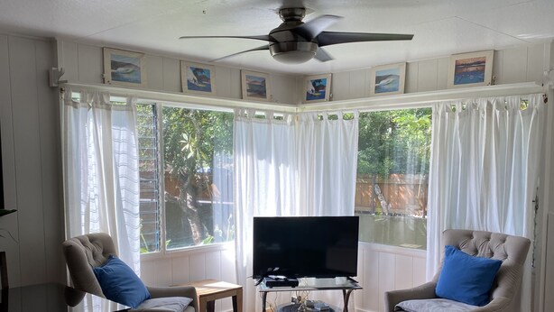 Uma sala de estar com ventilador de teto, dois poltronas, um sofá, uma televisão e uma janela com cortinas.