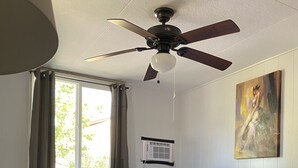 Quarto com cama, ventilador de teto, janela com cortinas, ar condicionado na parede e um quadro.