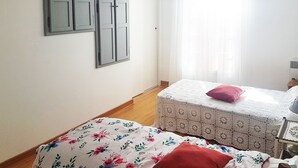 Ein Schlafzimmer mit Bett, einem kleinen Tisch und einem Sessel. Es gibt ein Fenster mit Vorhängen und ein an der Wand befestigtes Regal mit einem Spiegel.