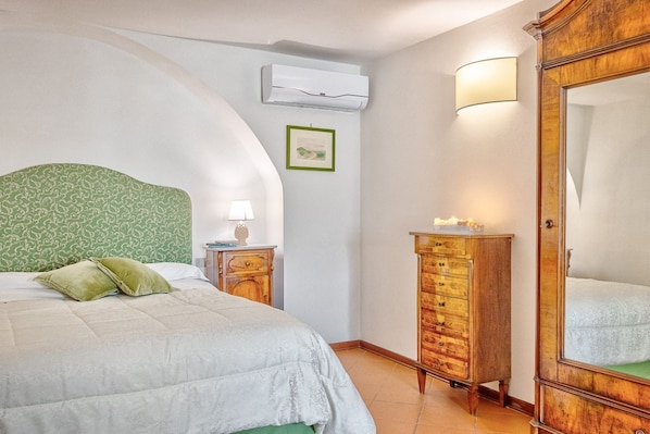 A bedroom with a curved headboard, a bed with green pillows, a bedside table with a lamp, a dresser, and a wall-mounted air conditioner.