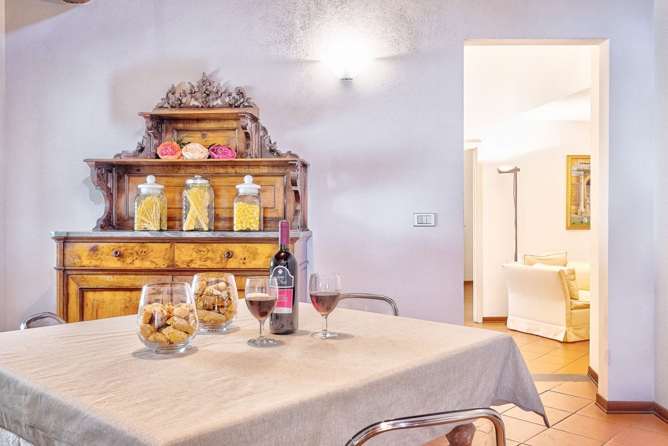 Un tavolo da pranzo con vasetti di vetro e bicchieri da vino, un mobiletto in legno con oggetti decorativi, e una porta che conduce in un'altra stanza.