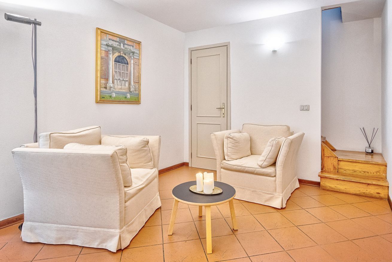 A room with two beige armchairs, a small round table with candles, a painting on the wall, a door, and a staircase.