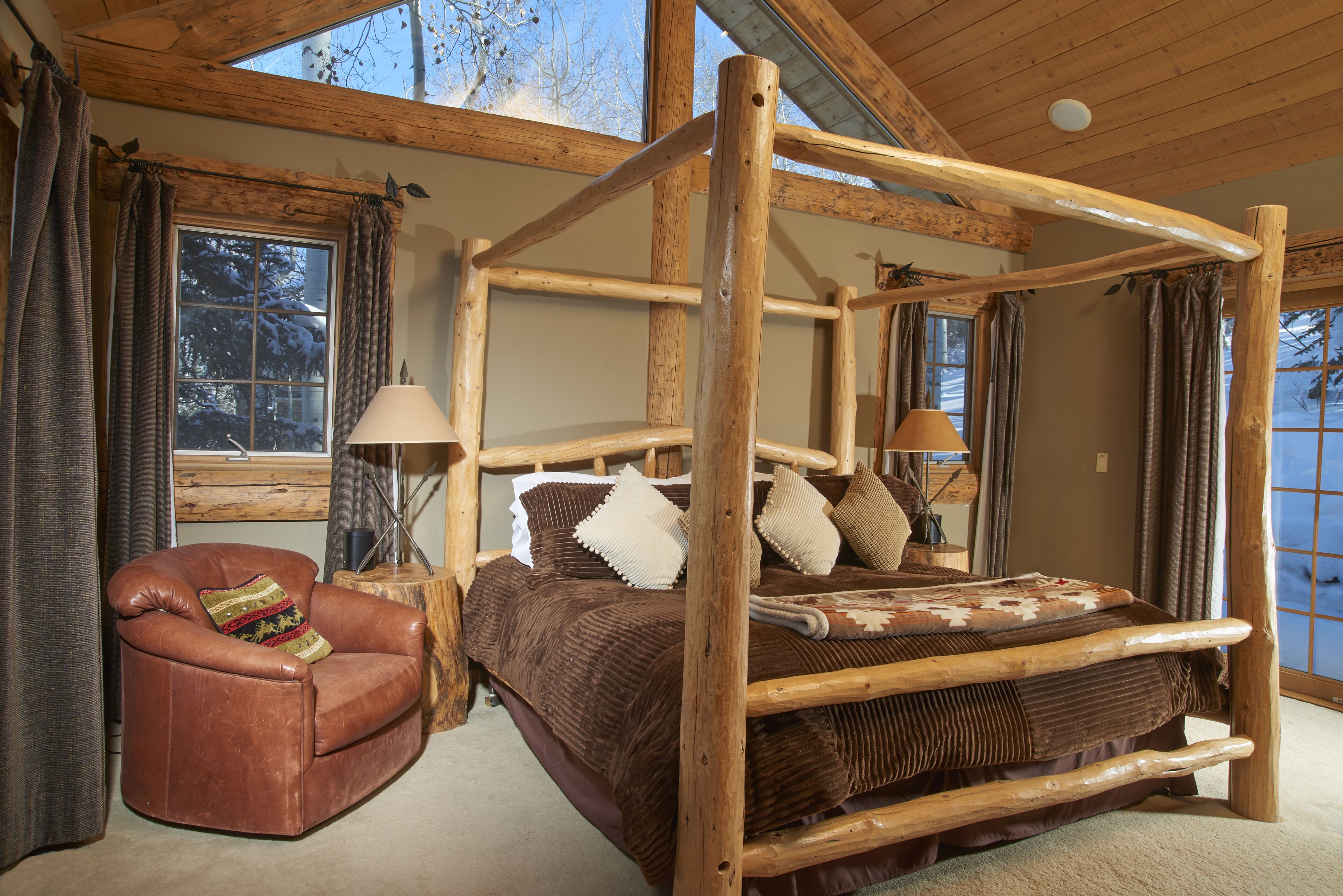 A log cabin bedroom with a four-poster bed, a leather armchair, and large windows.