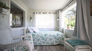 Un dormitorio con cama, dos sillas de mimbre, ventana con cortinas y vista a la vegetación.