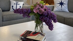 Una sala de estar con una mesa de centro blanca, una estantería llena de libros, un adorno con forma de velero y un sofá gris con almohadones de temática náutica.