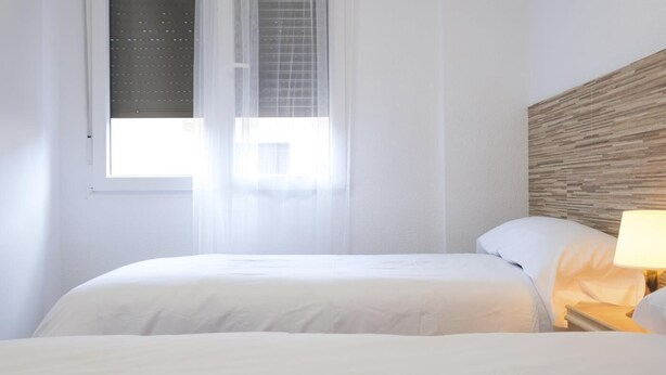 Een hotelkamer met een bed, nachtkastje met lamp en raam met dunne gordijnen.