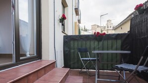 Een klein terras buiten, met een tafel en twee stoelen, een groene omheining en een raam met gordijn.