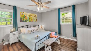 A bedroom with a bed, a ceiling fan, a nightstand, a bench, a TV, and a window with curtains. - Bright & Cheery Bungalow - Mins to Downtwn Fort Mill (Fort Mill)