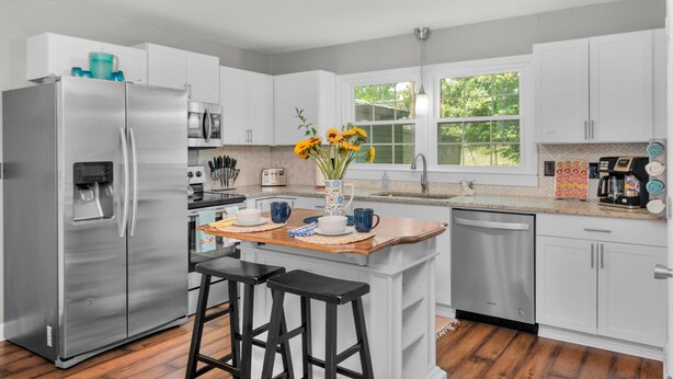 A modern kitchen with white cabinets, stainless steel appliances, a wooden island with stools, and a window providing natural light. - Bright & Cheery Bungalow - Mins to Downtwn Fort Mill (Fort Mill)