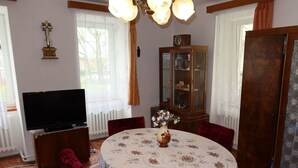 Un coin repas avec une table ronde, des chaises en velours rouge, un vase avec des fleurs, un téléviseur posé sur un meuble et un buffet en bois avec des portes en verre.