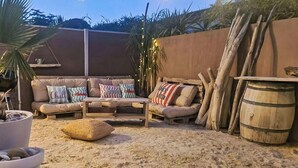 Ein sandiger Außenbereich mit einer Sofagarnitur, einem Couchtisch, einem Blumentopf und einem Holzfass.