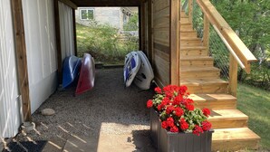 Een overdekt buitengebied met een houten trap die leidt naar een houten terras. Kajaks en een kanoan staan tegen de muur aangezet.