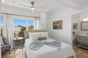 Un dormitorio con cama, mesita de noche, silla, ventana con cortinas, ventilador de techo y una obra de arte en la pared.