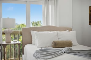 A hotel room with a bed, a bedside table with a lamp, and a view of greenery through the window.