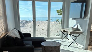 Ett modernt vardagsrum med en stor skjutdörr i glas som leder ut till en terrass med utsikt över stranden, en svart soffa, en vit fåtölj, en rund pall och ett litet bord med en korg.