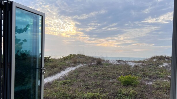Ett modernt rum med en glasdörr som leder ut till en terrass med utsikt över en strand med sanddyner och en molnig himmel.