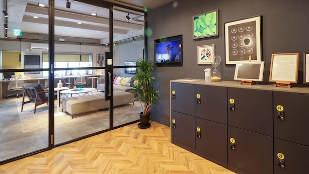 A modern office space with a large glass partition, a herringbone wood floor, a reception area with a plant, and a wall-mounted TV. - Deigo Hotel (Okinawa)