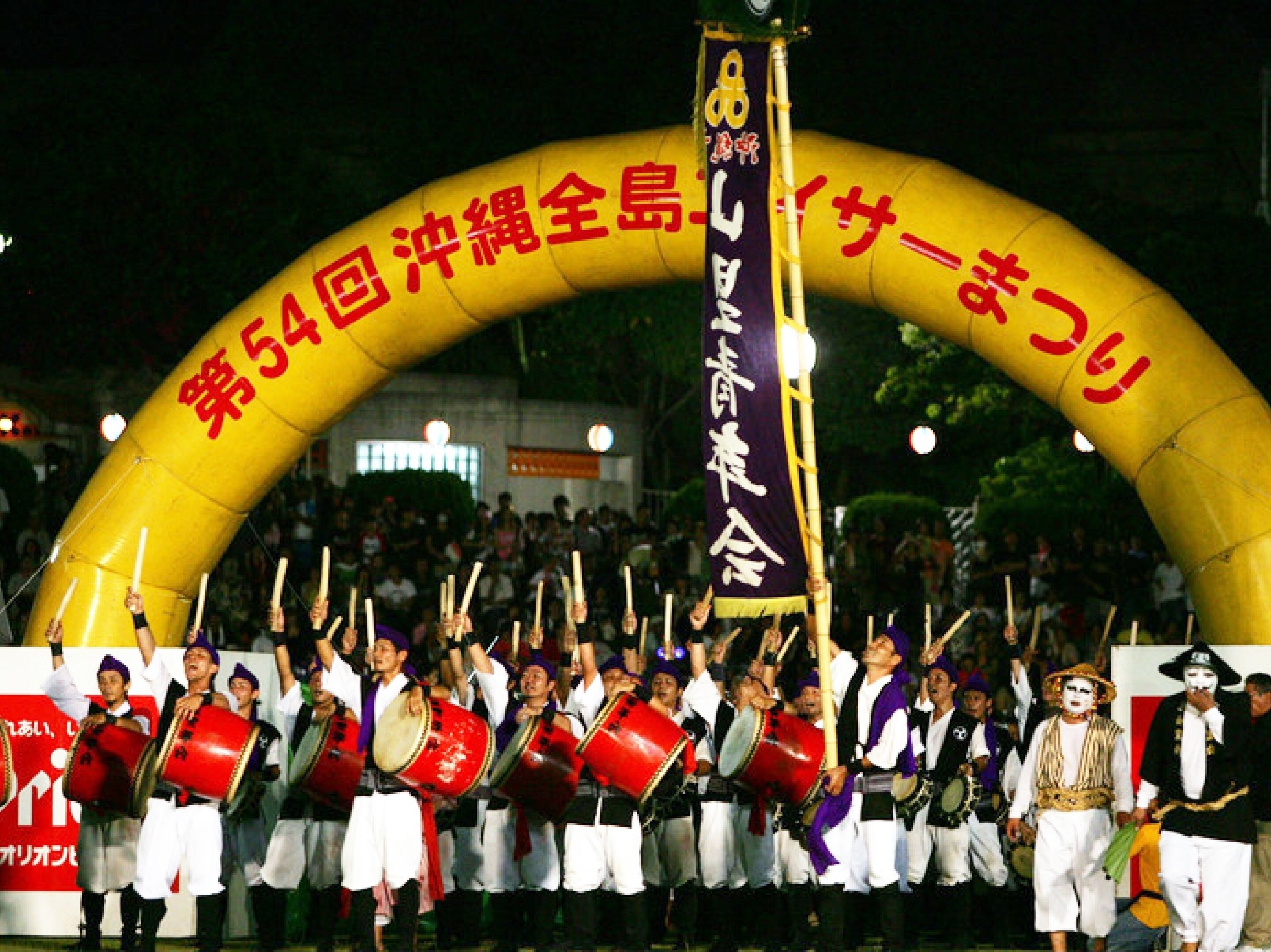 A group of people in traditional attire performing under a large arch with Japanese text.