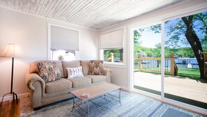 Ein Wohnzimmer mit einem beigefarbenen Sofa, gemusterten Kissen, einem Metall-Couchtisch und Blick auf eine Terrasse und Grünflächen.