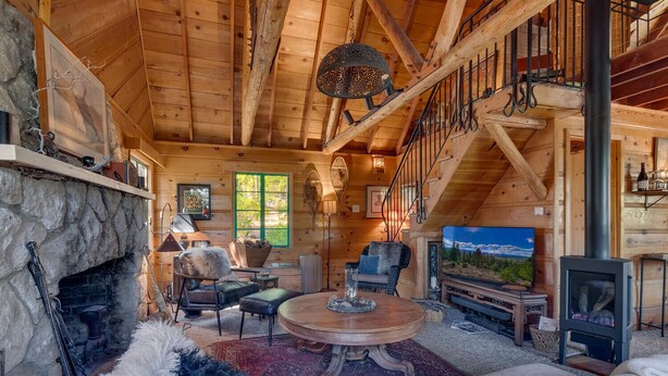 Ein gemütliches Blockhaus-Interieur mit Kamin aus Stein, Holzmöbeln, einem großen Fernseher und einer Treppe, die zu einer oberen Ebene führt.