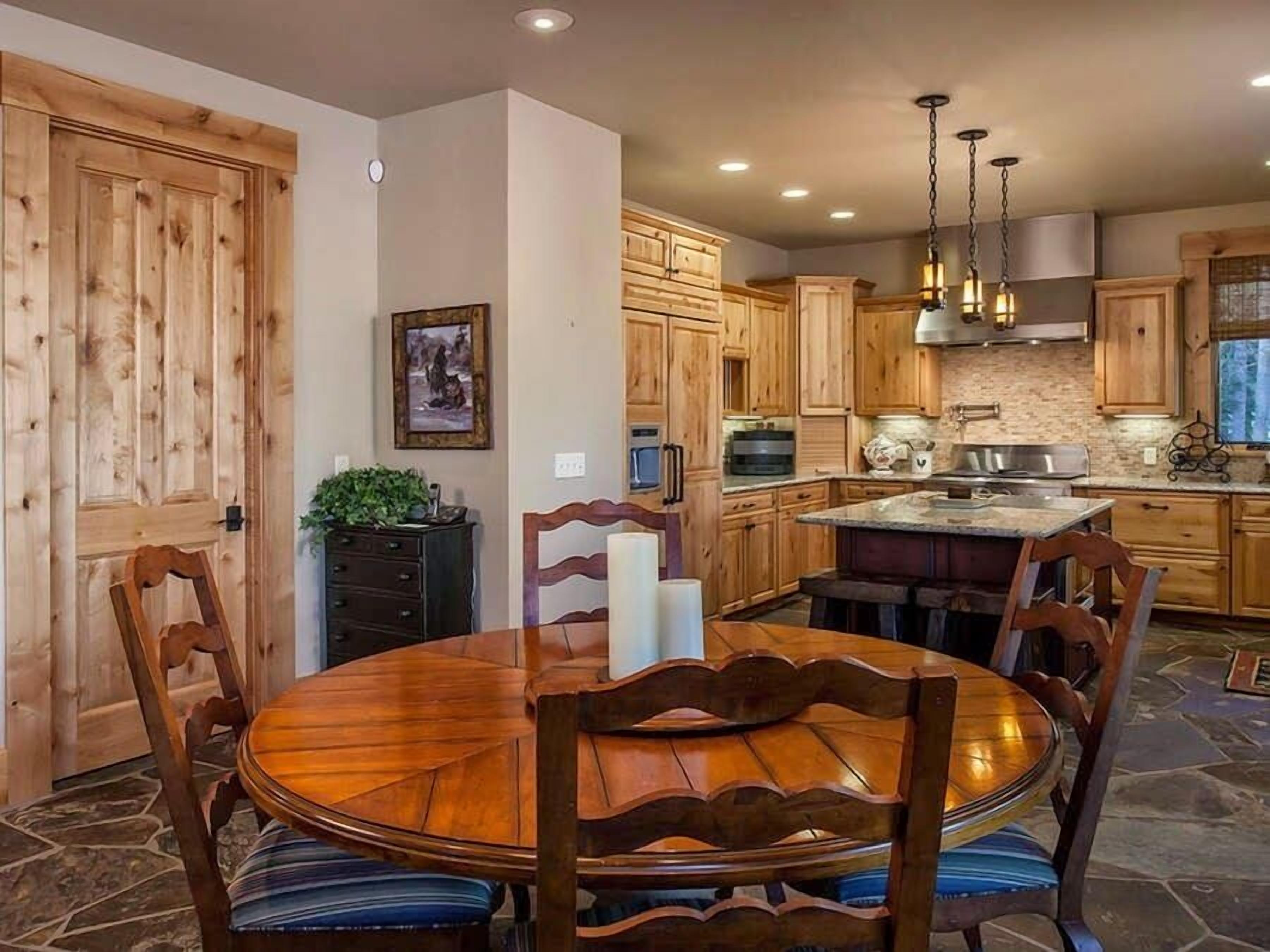 Una mesa de comedor de madera con sillas, una cocina con armarios de madera, piso de piedra y una lámpara colgante.