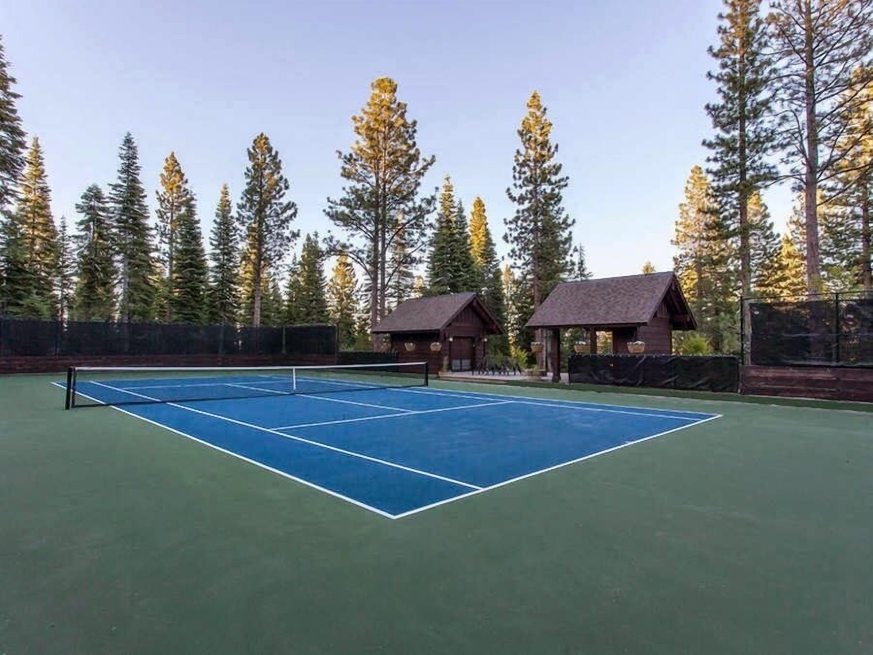 A tennis court with a net, surrounded by tall trees and two wooden structures.