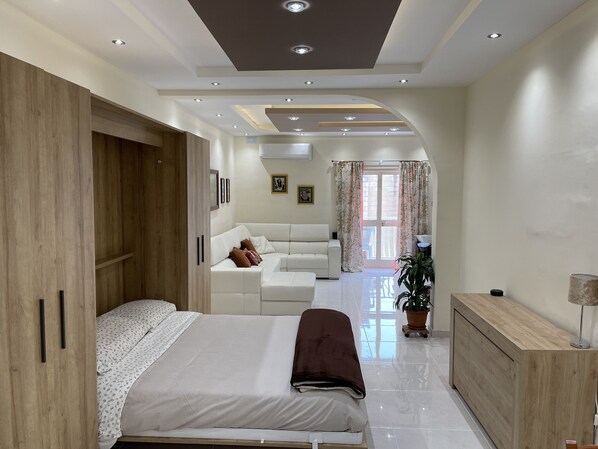 A modern bedroom with a large bed, a wooden wardrobe, a white sofa, and a small table with a lamp.