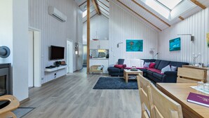 A spacious living area with a high ceiling, skylight, a flat-screen TV, a sofa, a dining table, and a fireplace.