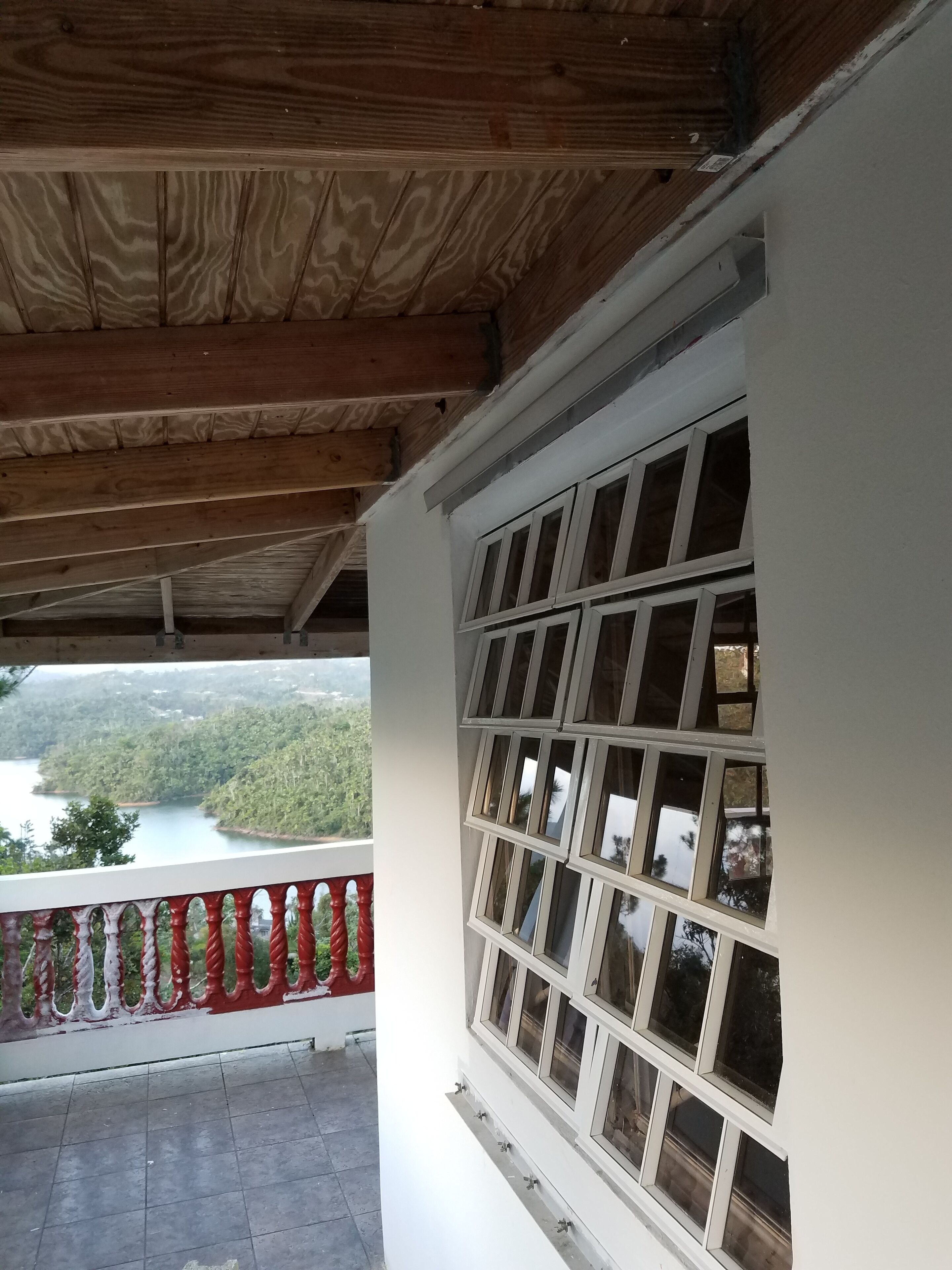 Un balcón con vistas a un lago y un bosque, vigas de madera y una pared blanca con varias ventanas.
