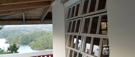 Un balcón con vistas a un lago y un bosque, vigas de madera y una pared blanca con varias ventanas.