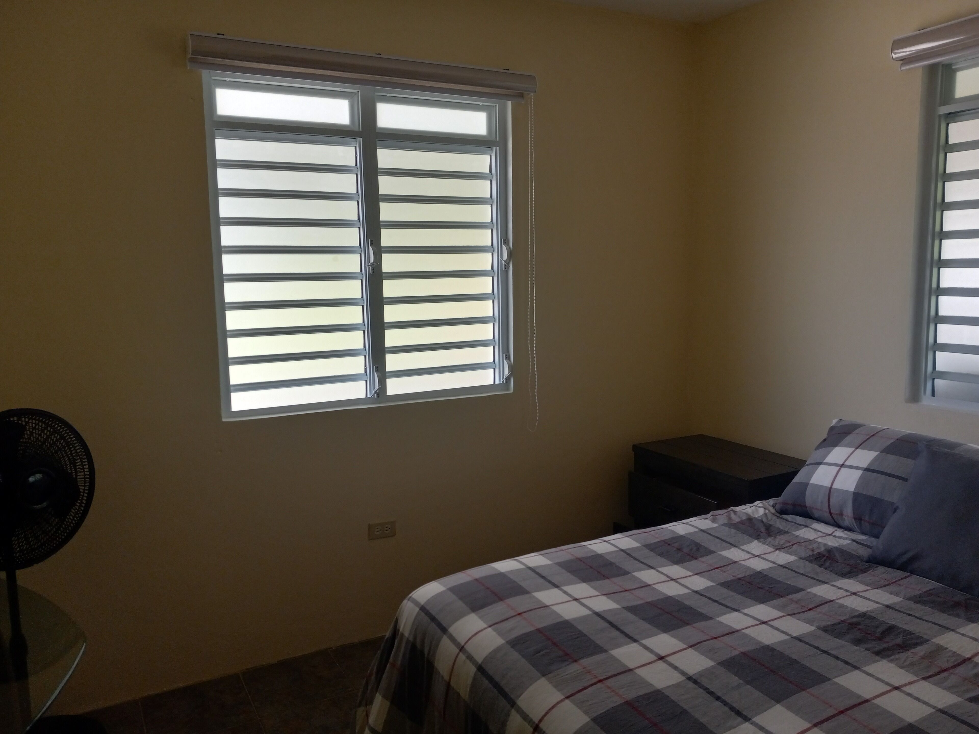 Un dormitorio con una cama, una mesita de noche, un ventilador y una ventana con persianas.