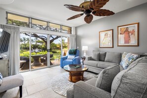 A spacious living room with a gray sofa, a blue chair, a glass coffee table, and a ceiling fan. There are large windows with a view of a garden.