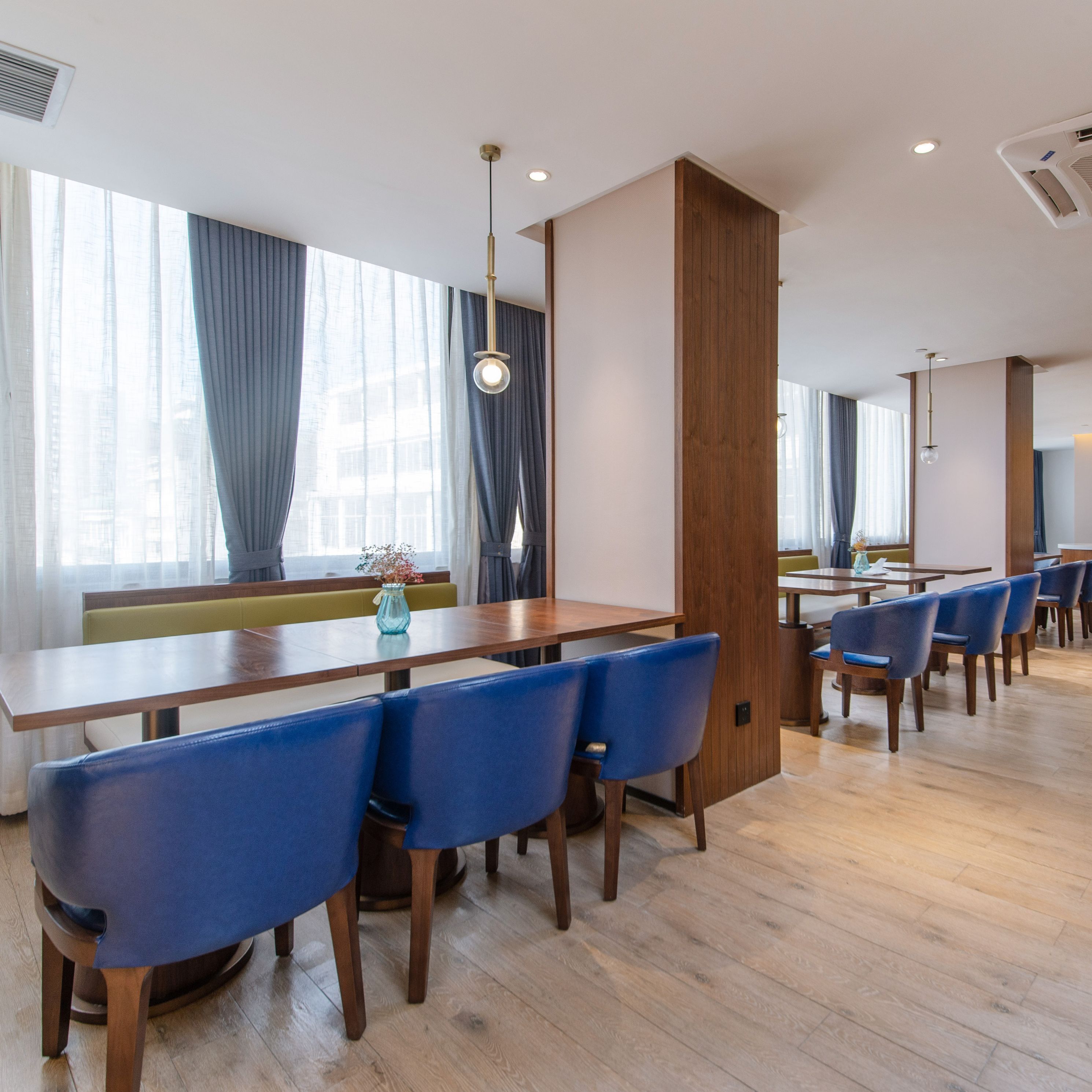 A modern dining area with wooden tables and blue chairs, large windows with curtains, and recessed ceiling lighting.