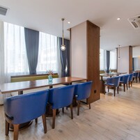 Un comedor moderno con mesas de madera y sillas azules, ventanas grandes con cortinas e iluminación empotrada en el techo.