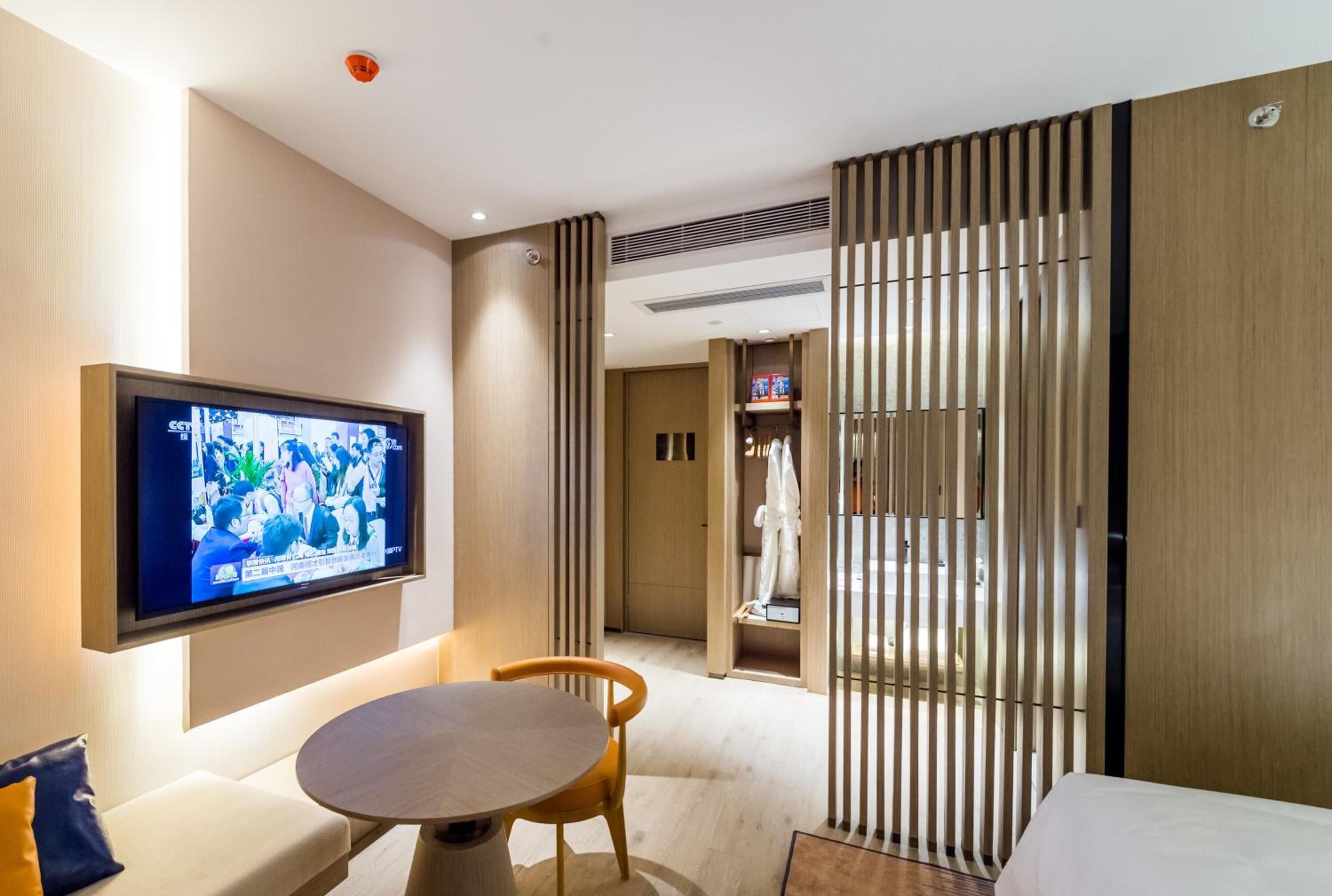 Una habitación de hotel moderna con un televisor de pantalla plana montado en la pared, una mesa redonda de madera, un sofá y una puerta corredera que da al baño.