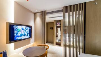 Una habitación de hotel moderna con un televisor de pantalla plana montado en la pared, una mesa redonda de madera, un sofá y una puerta corredera que da al baño.