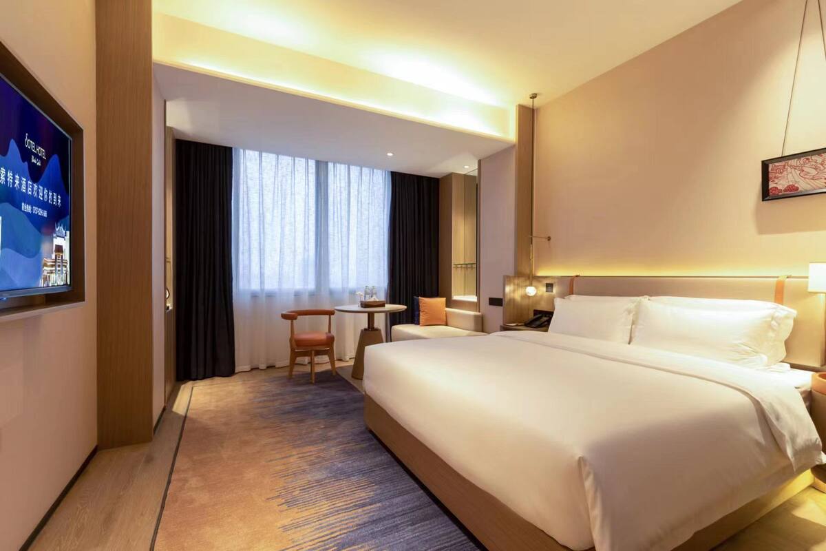 Una habitación de hotel con una cama grande, un escritorio con silla, un televisor en la pared y un ventanal con cortinas.